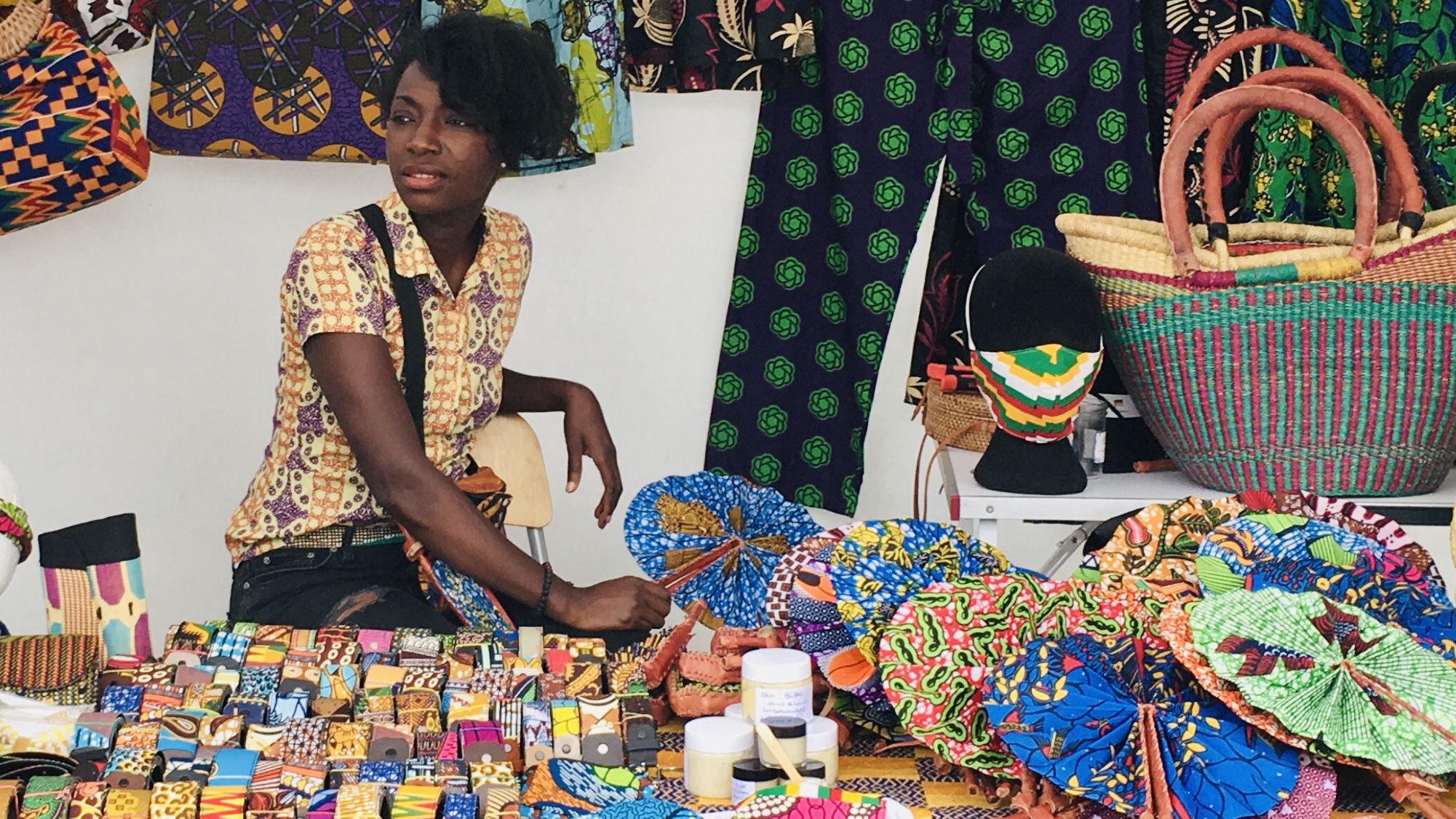 Regina Serwaa Dwomoh, Gründerin von MEDAASE, einem Shop für handgemachte Produkte aus afrikanischen Stoffen, sitzt aufmerksam an einem Marktstand mit ihren Textilien und Accessoires